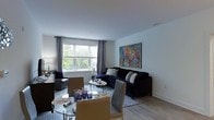 This well-appointed single-floor residence showcases a thoughtfully designed open-concept layout that seamlessly blends the dining and living areas into one cohesive, inviting space. The living area is anchored by a deep navy blue sofa adorned with decorative pillows, positioned beneath a large, vibrant abstract painting that serves as a bold focal point on the crisp white wall. A glass-top coffee table adds a contemporary touch, while a patterned area rug grounds the seating arrangement with subtle texture and elegance. The dining area features a sleek glass-top table surrounded by tan upholstered chairs, creating a sophisticated setting ideal for entertaining. Light hardwood-style flooring runs throughout, lending warmth and continuity to the space. A generous window dressed with grey curtains floods the interior with natural light and offers a view of lush greenery outside, enhancing the airy ambiance. Clean white walls and smooth ceilings contribute to a sense of openness, while carefully selected lighting fixtures and tasteful décor complete the polished, modern aesthetic. The overall atmosphere is refined yet comfortable, reflecting a balance of style and livability.