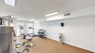 Located at 101 Meadowood St in Greensboro, NC, this fitness center offers a dedicated exercise space designed to support a well-rounded workout routine. The room features neutral white walls, a textured ceiling, and durable carpeted flooring that spans the entire area, creating a clean and functional environment.

The space is outfitted with a diverse selection of equipment to accommodate both cardio and strength training needs. Cardio offerings include a SportsArt treadmill and an elliptical machine, while strength training is supported by a multi-function cable and weight machine, a leg press/extension machine, and an adjustable weight bench. A three-tier dumbbell rack stocked with a full range of hex dumbbells provides additional free weight options.

Lighting is provided by recessed flush-mount fluorescent ceiling fixtures, ensuring bright, even illumination throughout. Large windows along one wall invite natural light into the room and offer views of an outdoor pool area, adding a pleasant visual connection to the surrounding grounds. A wall-mounted paper towel or sanitation dispenser and a stainless steel trash receptacle are conveniently placed for user hygiene and cleanliness. The space is accessed through a set of glass-panel doors, and a fire extinguisher is mounted near the entrance for safety.