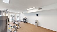 Located at 101 Meadowood St in Greensboro, NC, this fitness center offers a dedicated exercise space designed to support a well-rounded workout routine. The room features neutral white walls, a textured ceiling, and durable carpeted flooring that spans the entire area, creating a clean and functional environment.

The space is outfitted with a diverse selection of equipment to accommodate both cardio and strength training needs. Cardio offerings include a SportsArt treadmill and an elliptical machine, while strength training is supported by a multi-function cable and weight machine, a leg press/extension machine, and an adjustable weight bench. A three-tier dumbbell rack stocked with a full range of hex dumbbells provides additional free weight options.

Lighting is provided by recessed flush-mount fluorescent ceiling fixtures, ensuring bright, even illumination throughout. Large windows along one wall invite natural light into the room and offer views of an outdoor pool area, adding a pleasant visual connection to the surrounding grounds. A wall-mounted paper towel or sanitation dispenser and a stainless steel trash receptacle are conveniently placed for user hygiene and cleanliness. The space is accessed through a set of glass-panel doors, and a fire extinguisher is mounted near the entrance for safety.