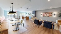This thoughtfully designed single-floor communal space offers a welcoming and versatile environment across two distinct zones. The open-plan layout is anchored by warm hardwood flooring throughout, recessed lighting, and a soft, light blue color palette that lends a calm, contemporary ambiance.

The kitchen area is a standout feature, showcasing bold blue cabinetry paired with a vibrant geometric triangle-patterned tile backsplash. A large butcher-block island with leather-seated bar stools serves as a central gathering point, complemented by a stainless steel refrigerator with ice dispenser, a built-in microwave, and a sink with open shelving displaying potted plants and dishware. Three striking black hourglass pendant lights hang above the island, adding an elevated design touch.

The lounge area offers a curated mix of seating, including a caramel leather sofa, deep navy accent chairs, and small café-style tables arranged for casual socializing. A vibrant floral artwork anchors one wall, while textured wallcovering and wall sconces add depth and warmth. A built-in banquette with terracotta upholstery and a herringbone-tiled floor accent provides an intimate nook beside sliding glass doors that open to an outdoor terrace, flooding the space with natural light and greenery views.
