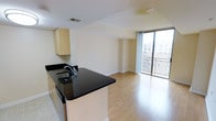 This single-floor space showcases a thoughtfully designed open-concept layout that seamlessly connects the kitchen and living area into one cohesive, light-filled environment. The kitchen is appointed with dark granite countertops that contrast elegantly against light cabinetry, and features a central island with a built-in sink — ideal for both meal preparation and casual entertaining. The flooring transitions from tile in the kitchen to warm hardwood-style planks throughout the living area, adding a sense of continuity and warmth. A standout feature of the space is the expansive floor-to-ceiling glass wall that floods the interior with abundant natural light and opens directly to a private balcony, offering open views of the surrounding cityscape. The neutral tones of the walls and cabinetry create a clean, versatile backdrop, while the open layout maximizes the sense of space and flow. Ceiling-integrated ventilation and smoke detection fixtures reflect practical, modern construction. The overall ambiance is bright, airy, and contemporary, making this a well-appointed space suited for comfortable everyday living.