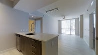 This single-floor space showcases a contemporary open-concept design that seamlessly connects the kitchen and living areas. The kitchen is anchored by a large island featuring a light quartz countertop, dark wood cabinetry with sleek bar-pull hardware, and an undermount sink paired with an industrial-style pull-down faucet. Directional track lighting overhead highlights the kitchen workspace, while a recessed light adds definition to the adjacent entry alcove, which features a warm yellow accent wall and a frosted glass door.

The living area extends naturally from the kitchen, finished with light-toned hardwood flooring that lends warmth throughout. A ceiling fan with a light fixture provides both comfort and ambient illumination. Floor-to-ceiling windows with horizontal blinds span the far wall, allowing abundant natural light to fill the space and offering views to the exterior. The neutral white and grey walls throughout create a clean, modern backdrop that complements the contemporary finishes. The open layout and thoughtful design elements combine to create a bright, functional, and inviting living environment.