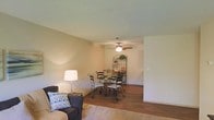 This single-story space presents an open-concept layout that seamlessly connects the living and dining areas, creating a cohesive and functional environment. The living area features warm hardwood flooring that extends into the dining space, lending a consistent and inviting aesthetic throughout. A plush area rug anchors the seating area, where a cushioned armchair with a decorative pillow provides a comfortable spot for relaxation. A table lamp adds a soft, warm glow to the corner.

The dining area is centered around a round glass-top table with ornate wrought iron chairs, contributing a classic and elegant touch to the space. A decorative wrought iron shelving unit against the wall provides both display and storage functionality, adding visual interest to the room.

The ceiling features a textured finish and is equipped with a ceiling fan fitted with a light fixture, offering both airflow and ambient lighting. Neutral-toned walls throughout the space create a calm and versatile backdrop, suitable for a variety of décor styles.

A connecting hallway or room is visible beyond the dining area, suggesting additional space within the unit. Overall, the space conveys a warm and practical atmosphere with simple, comfortable furnishings and classic design elements.