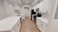 This single-floor space showcases a thoughtfully designed galley-style kitchen as its centerpiece. The kitchen features crisp white shaker cabinets lining both walls, extending to the ceiling to maximize storage capacity. Sleek bar-pull hardware in brushed silver adds a contemporary touch throughout. Light-toned luxury vinyl plank flooring runs the length of the kitchen, providing both durability and a warm aesthetic contrast to the white cabinetry.

The countertops are finished in a clean white stone material, offering generous prep space along both sides of the galley layout. A stainless steel undermount sink with a modern faucet is positioned along the left wall, accompanied by a built-in dishwasher. On the right side, a black gas range with stainless steel accents serves as the cooking focal point, complemented by an overhead range hood. A black refrigerator anchors the far end of the kitchen.

Recessed lighting illuminates the space evenly from above, while a defined beam detail adds subtle architectural interest to the ceiling. A hallway visible at the far end of the kitchen connects to the remainder of the unit, suggesting additional living space beyond.