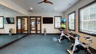 Located at 1951 Old Haywood Rd in Asheville, NC, this single-floor fitness studio is thoughtfully designed to support a variety of workout styles. The space is divided into distinct functional zones, seamlessly connected by consistent design elements throughout.

The main exercise area features patterned teal carpet flooring and is equipped with spinning bikes, a BOSU balance trainer, and a tiered rack of colorful exercise balls. A wall-mounted flat-screen television and multiple windows with blinds allow for natural light from the surrounding greenery, creating an energizing atmosphere. A ceiling fan overhead aids in air circulation during workouts.

One wall is fully covered by floor-to-ceiling mirrors, dramatically expanding the sense of space while allowing users to monitor their form. Adjacent to this is a striking accent wall clad in warm wood planking, adding a natural, grounding contrast to the otherwise modern aesthetic. Wall-mounted racks hold rolled yoga mats, making them easily accessible for stretching or studio-style classes.

Recessed lighting and a clean, neutral color palette keep the environment bright and focused. Glass-panel doors connect the studio to additional areas, reinforcing the open, welcoming character of this well-appointed fitness facility.