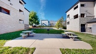 Located at 1435 W Talmage St in Springfield, Missouri, this outdoor common area is a well-maintained exterior space designed for community gathering and leisure. The centerpiece of the grounds is a neatly landscaped courtyard nestled between multi-story residential buildings clad in a combination of white siding and stone veneer exteriors. The courtyard features a broad concrete patio bordered by lush green lawn, providing a clean and functional gathering space. Several concrete picnic tables with attached benches are positioned throughout, offering ample seating for outdoor dining or socializing. A communal grill station is conveniently situated on the patio, making the space well-suited for outdoor cookouts. Mature trees and decorative rock landscaping add natural character to the grounds, while a pathway connects the courtyard to the surrounding buildings. The property includes a generously sized parking lot along the perimeter, accommodating multiple vehicles with clearly marked spaces and easy access via sidewalks. The overall setting is open and bright, benefiting from abundant natural light and open sky views. The grounds are well-kept and thoughtfully arranged to serve as a functional and inviting outdoor amenity for residents of the community.