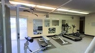 This single-floor fitness room offers a practical and functional workout environment designed to accommodate a range of exercise needs. The space is outfitted with a selection of cardio equipment, including a treadmill and an elliptical machine, positioned along one wall to maximize floor space. Adjustable weight benches and a dumbbell rack provide options for strength training, while a cable machine adds versatility for resistance exercises. Rubber floor mats are placed beneath the equipment, offering protection and stability during workouts. Large wall-mounted mirrors span a significant portion of one wall, visually expanding the room and allowing users to monitor their form. Fluorescent strip lighting runs across the textured ceiling, ensuring the space is well-lit throughout. A window on one wall introduces a degree of natural light and a connection to the outdoors. The walls are finished in a neutral tone, contributing to a clean, no-frills atmosphere. Overall, the space is efficiently organized to deliver a straightforward and accessible fitness experience.