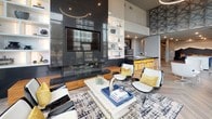This contemporary amenity lounge is designed to impress, offering a seamlessly connected single-floor layout that balances elegant aesthetics with functional comfort. The space is anchored by dramatic floor-to-ceiling windows that flood the interior with natural light while framing views of the surrounding outdoor terrace and cityscape beyond. Herringbone-patterned flooring in a warm wood tone flows throughout, lending a refined yet inviting character to the entire space.

The main lounge area features a striking dark slab fireplace wall with an integrated flat-screen television, flanked by open display shelving and surrounded by a curated mix of modern seating, including sleek accent chairs and a geometric patterned area rug. Adjacent to the lounge, a fully equipped wet bar offers wine storage, beverage stations, and stylish cabinetry with overhead glass rack displays, ideal for entertaining.

A sunlit seating nook near the expansive windows provides an intimate gathering spot, furnished with sculptural contemporary chairs and small tables. A separate built-in banquette area showcases a standout textured wall mural depicting a mountain landscape with dimensional bird silhouettes, creating a dramatic and artistic focal point. Recessed lighting and bold decorative accents in navy, gold, and yellow complete this thoughtfully designed communal destination.