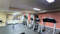 This fitness center is designed to accommodate a variety of workout routines within a functional, single-floor layout. The space features warm terracotta-toned accent walls complemented by neutral tiling and light-colored ceilings, creating an energizing yet approachable atmosphere. Overhead fluorescent lighting ensures the entire area is well-illuminated throughout the day.

The cardio section is prominently equipped with an elliptical trainer, a stationary bike, and additional aerobic machines visible toward the rear of the room. A dedicated free weights area includes a multi-tiered dumbbell rack stocked with a full range of weights, alongside a workout bench for strength training exercises. Large wall-to-wall mirrors along one side visually expand the space and allow users to monitor their form during workouts.

The flooring throughout is a durable, easy-to-maintain tile suitable for high-traffic fitness use. The layout is thoughtfully organized to allow clear circulation between equipment zones. Overall, the fitness center offers a practical and well-maintained environment suited for both cardiovascular and strength training activities.