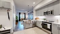 This thoughtfully designed 1-bedroom, 1-bathroom residence offers a cohesive blend of modern aesthetics and functional living. The open-concept layout connects the kitchen and living room seamlessly, creating an inviting and efficient use of space. The kitchen is a standout feature, showcasing gray shaker cabinetry, a white hexagonal tile backsplash, stainless steel appliances including a range, dishwasher, microwave, and refrigerator, along with a peninsula island with a farmhouse-style sink and butcher block countertop — ideal for casual dining with bar stool seating beneath pendant lighting. Wood-look flooring flows throughout the main living areas, complemented by recessed lighting and a decorative mid-century modern ceiling fixture in the living room, which also benefits from natural light through large windows dressed with dark drapery panels. The bedroom offers a serene retreat with a distinctive wood-paneled accent wall, plush carpeting, and generous window coverage. The bathroom features gray cabinetry, a white quartz countertop, an illuminated mirror, and a separate water closet. A dedicated closet provides organized storage. The overall ambiance is modern, warm, and polished.