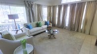 This single-floor living space showcases a thoughtfully designed interior that balances elegance with comfort. The living area is anchored by a crisp white sofa adorned with vibrant accent pillows in shades of teal, blue, and green, creating a lively yet refined color palette against the neutral, carpet-covered floor. A round glass-top coffee table with a gold cross-base serves as a central focal point, topped with a decorative floral arrangement that adds a natural touch to the room.

Floor-to-ceiling drapery in a muted taupe-gray tone frames the multiple large windows that line the walls, allowing generous natural light to fill the space while maintaining an air of sophistication. The arched window detail visible above adds an architectural charm that elevates the overall aesthetic. Sheer panels layered beneath the drapes provide softened diffusion of light throughout the day.

The neutral wall tones and carpeted flooring create a warm, cohesive backdrop that complements the carefully curated furnishings. Additional seating and accent tables are positioned to encourage conversation, making this a welcoming and functional living environment suited for both relaxed everyday living and entertaining.
