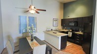 This single-story space showcases an open-concept design that combines the kitchen and dining areas into one cohesive living environment. The most striking architectural feature is the dramatically high ceiling, which creates an impressive sense of volume and airiness throughout the space. A ceiling fan is centrally positioned overhead, adding both functionality and visual interest to the room.

The kitchen is equipped with dark espresso-toned cabinetry that provides a bold contrast against the lighter walls and countertops. Black appliances, including a range and microwave, complement the cabinetry's rich tones. A peninsula counter extends outward, creating a natural division between the kitchen and dining areas while also offering additional workspace and a casual seating option. The sink is integrated into the peninsula, adding practicality to the layout.

The dining area accommodates a full-sized table and sits adjacent to the kitchen, making it ideal for everyday meals or entertaining. An accent wall in a muted sage green adds a layer of depth and color to the space. The flooring transitions between hardwood-style and carpeted surfaces, subtly defining each zone. Artwork and decorative elements throughout contribute to a warm, welcoming ambiance.