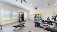 This fitness center is a thoughtfully designed, single-room workout facility that provides a comprehensive range of exercise equipment within a compact yet efficient footprint. The space features durable speckled rubber flooring throughout, offering both traction and comfort during workouts, while crisp white walls and recessed lighting create a clean, energizing atmosphere. Large windows fitted with blinds line the walls, inviting ample natural light and contributing to an open, airy feel.

The cardio section is well-appointed with treadmills, an elliptical trainer, a stationary bike, and a rowing machine, catering to a variety of aerobic exercise preferences. For strength training, the facility includes a multi-functional cable machine, a two-tier dumbbell rack with a full range of free weights, a flat workout bench, and a dedicated rack stocked with medicine balls and kettlebells. A large wall mirror spans one side of the room, visually expanding the space while allowing users to monitor their form.

Overhead, multiple large ceiling fans ensure consistent airflow and comfort during intense sessions, complemented by a wall-mounted flat-screen television for entertainment. The overall ambiance is modern, bright, and functional — a well-rounded fitness space designed to support a full-body workout routine.