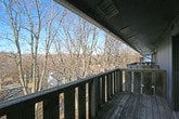 This single-floor space is distinguished by its generous outdoor deck, which serves as a standout architectural feature. Constructed with weathered wood planking and sturdy wooden railings, the deck extends along the exterior of the structure, offering an elongated outdoor living area that invites relaxation and open-air enjoyment. The deck is accessed via sliding glass doors fitted with vertical blinds, which allow for flexible light control while maintaining a visual connection between the interior and the outdoors.

Positioned at an elevated vantage point, the deck provides expansive views of a dense, mature woodland. Bare deciduous trees frame the horizon under open blue skies, creating a serene, nature-immersed atmosphere that shifts with the seasons. The surrounding landscape includes glimpses of neighboring structures nestled among the trees, adding a sense of community while preserving a feeling of natural seclusion.

Overhead, the deck is partially sheltered by the building's eave, offering some protection from the elements. Ventilation units are visible along the soffit, reflecting the practical infrastructure of the space. The blue-gray exterior siding of the building contrasts with the warm tones of the natural cedar wood accents on the deck enclosure. Overall, this space delivers a compelling blend of functional outdoor living and scenic natural surroundings.