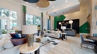 Located at 2900 University Square Drive in Tampa, Florida, The Standard at Tampa presents a thoughtfully designed single-floor clubhouse and leasing space that makes an immediate impression. Floor-to-ceiling glass entry doors flood the entryway with natural light, framed by a dramatic floor-to-ceiling wood accent wall adorned with a geometric arrow-pattern tile overlay in muted teal tones. This striking architectural feature anchors multiple seating vignettes, including a cozy lounge with a teal velvet sofa, sculptural wooden stools, and vibrant green abstract artwork. The expansive main lounge area features light wood-look tile flooring, soaring ceilings with oversized circular pendant acoustic panels in green, black, and natural wood tones, and generous seating arrangements with sofas, armchairs, and dining tables designed for both relaxation and socializing. Large windows along the perimeter invite views of the surrounding landscape and bathe the interior in warm Florida sunlight, complemented by rich emerald green drapery and bold green accent walls. A sleek, dark reception desk with a sculptural cluster pendant light serves as the hub for the leasing office area, with a glass-walled private office nearby. The overall ambiance balances contemporary design with inviting warmth, creating a vibrant communal environment ideal for the university community it serves.