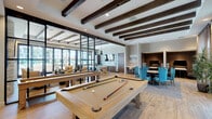This single-floor community amenity space is thoughtfully designed to blend comfort and functionality in a modern, upscale setting. Exposed dark wood ceiling beams run throughout, adding warmth and architectural character to the bright, open interior. Wood-look flooring flows seamlessly across the space, unifying the distinct activity zones.

The entry corridor welcomes visitors through sleek, floor-to-ceiling glass and dark metal-framed doors, opening into a spacious interior. A dedicated game area features a full-size billiards table as its centerpiece, positioned beneath the striking beamed ceiling. Adjacent to this is a well-appointed kitchen with a large white quartz island, dark wood cabinetry, stainless steel appliances including a double oven, and a bold geometric tile backsplash. Industrial-style pendant lights hang above the island, adding to the contemporary aesthetic.

The lounge area offers a comfortable gathering space anchored by a floor-to-ceiling stacked stone fireplace with a mounted flat-screen television above. Plush seating in warm gold and tan tones invites relaxation. Large sliding glass doors and expansive windows flood the space with natural light and provide direct views and access to an outdoor terrace and surrounding landscape, creating a seamless indoor-outdoor connection throughout.