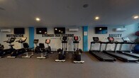 This fitness center is thoughtfully designed to support a comprehensive workout experience within a single, open floor layout. The space is defined by a striking color scheme of teal-blue accent columns and walls contrasted against grey tones, creating an energetic and focused atmosphere reinforced by bold motivational phrases such as "Stronger Than Yesterday," "Attitude," "Focus," and "Do It." displayed throughout.

The cardio zone is well-stocked with multiple treadmills, elliptical trainers, and stationary bikes, each equipped with individual digital displays. Multiple flat-screen televisions are mounted along the walls, keeping exercisers entertained during workouts. A dedicated strength and functional training area features a cable machine, adjustable weight bench, free weights, and a heavy punching bag on a freestanding rack, offering versatile training options.

A mirrored wall section expands the sense of space and aids in form correction during exercises. Stability balls, a BOSU trainer, and resistance accessories are neatly organized in a corner near a large window, which introduces natural light into the space. Recessed ceiling lighting and wall-mounted mini-split air conditioning units ensure a comfortable environment. The flooring transitions between carpet and rubber matting in functional training zones, enhancing safety and usability throughout.