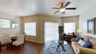This single-floor space is thoughtfully designed to feel open and welcoming. The living area immediately draws attention with its vaulted ceiling, which adds a sense of height and airiness to the room. A classic ceiling fan with a built-in light fixture serves as a functional centerpiece overhead, helping to circulate air throughout the space. Rich, warm-toned hardwood-style flooring runs throughout, providing a cohesive and polished foundation that complements the neutral, earth-toned walls. Natural light is a defining feature of this space, with multiple windows fitted with white horizontal blinds flanking a prominent sliding glass door dressed in vertical blinds. Together, these elements allow for generous daylight while maintaining privacy control. The sliding door offers a seamless connection to an outdoor area, enhancing the sense of openness. The living area comfortably accommodates a seating arrangement anchored by a patterned area rug in shades of blue and white, adding a decorative touch to the space. A glass-top coffee table and additional accent furniture contribute to a clean, casual aesthetic. Lush indoor plants and simple side furniture complete the inviting atmosphere, making this a comfortable and well-lit living environment.