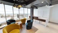 This contemporary single-floor space combines industrial and modern design elements to create a polished, welcoming environment. The open-plan lounge area is anchored by expansive floor-to-ceiling windows that flood the interior with natural light and offer sweeping panoramic views of the surrounding landscape, extending out to a railed outdoor terrace visible beyond the glass. The exposed black ductwork and industrial-style pendant lighting contrast stylishly against the white ceiling panels and light-toned tile flooring, giving the space a refined yet urban character. Structural concrete columns add an architectural authenticity to the design. The lounge is furnished with curved, pod-style chairs in bold mustard yellow and teal tones, arranged around natural wood stump accent tables, creating a vibrant and inviting seating area. Adjacent to the lounge, a sleek reception desk finished in rich dark wood provides a professional focal point, complemented by shuttered windows that offer a nod to classic design. The overall ambiance is bright, airy, and sophisticated — ideal for a collaborative or client-facing commercial environment that balances comfort with a strong design identity.