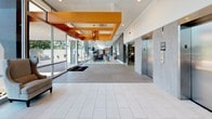 This single-floor lobby space presents a thoughtfully designed contemporary environment that balances functionality with striking visual appeal. The elongated layout is anchored by bold orange ceiling beams and coffers that define distinct zones throughout the space, creating a dynamic architectural framework that draws the eye from entry to interior.

Upon entry, large-format light gray porcelain tile flooring establishes a clean, polished foundation. A branded entryway mat leads guests into an open foyer, where a statement geometric chandelier in warm amber tones hangs beneath the vibrant orange ceiling detail. Stainless steel elevator doors, framed by gray stone-clad surrounds, are prominently positioned along one wall, providing a sleek, functional focal point.

The lounge area features comfortable gray upholstered sofas accented with blue and orange throw pillows, arranged atop layered area rugs. A textured dark brick accent wall adds depth and contrast, complemented by curated décor including round mirrors and accent lamps in matching orange tones. Lush potted plants introduce natural warmth throughout.

Floor-to-ceiling glass walls flood the space with abundant natural light and offer open views of the surrounding landscape, including mature trees and a decorative stone water feature just outside. The overall ambiance is modern, polished, and inviting.