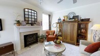 This single-floor space exudes warmth and character through its thoughtfully designed living area. The room centers around a classic tiled fireplace, framed by a white mantel adorned with decorative accents including candles and a striking multi-pane mirror. Crown molding runs along the ceiling, adding an elegant architectural touch throughout. Natural light filters in through a window dressed with patterned curtains, brightening the neutral, carpeted interior. A ceiling fan overhead ensures year-round comfort. The living area is furnished with a plush orange armchair, a round ornate coffee table, and a rich wooden armoire paired with an open display cabinet — offering both storage and a showcase for curated décor. Decorative elements such as a lantern, clock, woven baskets, and seasonal accents lend the space a warm, eclectic personality. A potted plant nestled beside the fireplace adds a touch of greenery. The overall ambiance is cozy and lived-in, blending traditional furnishings with decorative layering to create an inviting retreat that feels both functional and full of personal character.