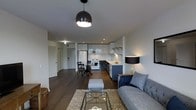This single-floor residence offers a thoughtfully designed open-concept layout that connects the living, dining, and kitchen areas into one cohesive and functional space. The living area is anchored by a tufted gray sofa, a glass-and-metal coffee table, and a plush area rug, creating a comfortable and stylish gathering space. A bold dome pendant light hangs overhead, adding a striking focal point to the room.

The kitchen is outfitted with sleek two-tone cabinetry — crisp white upper cabinets contrast with deep navy-blue lower units — paired with a white subway tile backsplash and light countertops for a clean, modern aesthetic. A white range, stainless steel microwave, and full-size refrigerator complete the kitchen's functional layout, while open shelving adds an airy, contemporary touch.

Adjacent to the kitchen, a compact dining nook with a round table and cross-back chairs provides an intimate space for meals. Dark hardwood flooring runs throughout the space, grounding the design and complementing the neutral white walls. Recessed lighting and carefully placed floor lamps ensure warm, layered illumination across the entire floor plan. An entryway with a door is visible in the background, completing the well-organized layout of this modern home.