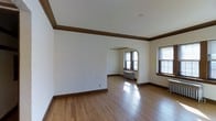 This single-floor space showcases a blend of classic architectural elements that give it a distinctly traditional character. The main living area is generously proportioned, with smooth white walls accented by rich, dark wood crown molding and matching baseboards that frame the room with a refined elegance. Hardwood flooring in a warm honey tone runs throughout, reflecting natural light and adding to the inviting atmosphere.

A prominent arched doorway serves as the defining architectural feature, connecting the main living area to an adjoining room and lending a sense of graceful continuity to the layout. This arched passage is complemented by the dark wood trim, reinforcing the cohesive traditional aesthetic throughout.

Natural light filters in generously through multiple windows fitted with dark wood frames, including an arched window that mirrors the doorway's design. A cast-iron radiator beneath one of the windows hints at the building's heritage and provides a period-appropriate detail. The ceilings are well-proportioned, contributing to an open, airy feel. Overall, the space presents a clean, well-maintained interior with strong architectural bones and classic detailing throughout.