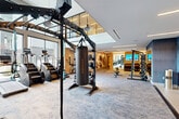 This expansive two-floor fitness center is designed to deliver a comprehensive workout experience in a thoughtfully appointed setting. The main floor commands attention with soaring floor-to-ceiling windows that flood the space with natural light and offer open views of the surrounding neighborhood. Pendant lighting and warm wood accents complement the modern aesthetic, while patterned carpet flooring provides a refined backdrop for a full suite of cardio equipment, including treadmills, stair climbers, and stationary bikes, all positioned to take advantage of the panoramic views. A prominent functional training rig anchors the main floor, complete with battle ropes, a heavy bag, and cable attachments for versatile strength training. A dedicated strength area features barbell racks, weight benches, a leg press, and an extensive dumbbell and kettlebell collection displayed on tiered racks alongside floor-to-ceiling mirrors. A glass-railed wood staircase with built-in lockers and cubby storage connects the floors with an elegant transition. The upper level houses a bright studio with light hardwood floors, a distinctive wave-form acoustic ceiling installation, large mirrors, stability balls, and open space for group or individual training sessions. Artistic wall murals add a creative energy throughout, making this facility as visually engaging as it is functionally complete.