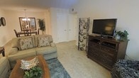 This single-floor residential space offers a comfortable and well-appointed living environment designed with practicality and warmth in mind. The living area serves as the central gathering space, featuring plush wall-to-wall carpeting in a neutral beige tone that adds softness underfoot and contributes to the room's cozy ambiance. A patterned area rug in muted blue and grey tones anchors the seating arrangement, which includes a cushioned sofa adorned with decorative throw pillows in complementary earth tones. A rich, dark-wood media console houses a flat-screen television, providing a natural focal point for the room, while a decorative metal panel adds an artistic accent nearby. A wooden coffee table grounds the seating area, and lush potted greenery brings a touch of nature indoors, brightening the space alongside framed wall art. The walls are finished in a soft, warm beige that enhances the welcoming atmosphere throughout. Two white interior doors along the back wall lead to additional areas of the unit, suggesting well-defined private spaces. Overhead ventilation is visible, reflecting thoughtful utility integration. Overall, the space presents a functional, comfortable layout with a cohesive and understated aesthetic.