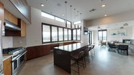 Located at 624 Yale Avenue North in Seattle's South Lake Union neighborhood, this single-floor communal space combines modern design with high-end functionality. The centerpiece is a spacious, fully equipped chef's kitchen featuring warm wood cabinetry, stainless steel appliances including a professional-grade range, and a large island with a dark countertop and bar seating. Pendant lighting and clerestory windows above the cabinetry flood the kitchen with natural light while framing panoramic views of the Seattle skyline and surrounding cityscape. Polished concrete floors run throughout, lending a sleek, cohesive feel to the entire space. Adjacent to the kitchen, a flexible lounge area offers a variety of seating arrangements, including patterned accent chairs gathered around a gold-topped circular table, built-in upholstered banquette seating, and intimate conversation groupings with woven accent chairs. Floor-to-ceiling windows and glass doors open the lounge to abundant natural light and outdoor views of the waterfront and city beyond. The interplay of natural wood accents, crisp white walls, and contemporary furnishings creates an inviting, upscale atmosphere well-suited for social gatherings and collaborative use.