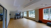 Located at 1 Kennedy Ave in Danbury, Connecticut, this single-floor fitness studio is designed to support an active and structured workout experience. The space centers around a dedicated cycling studio, thoughtfully laid out with multiple stationary bikes arranged to face a large wall-mounted flat-screen television — ideal for instructor-led or on-demand cycling sessions. The room features smooth, light-toned hardwood-style flooring that provides both durability and a clean, modern aesthetic. Neutral toned walls are accented by a bold orange column detail, adding a dynamic visual element to the space. Recessed lighting in the dropped ceiling ensures bright, even illumination throughout, complemented by an abundance of natural light streaming in through a row of large windows along the exterior wall. These windows also offer pleasant views of the surrounding outdoor greenery, creating an open and energizing atmosphere. The ceiling incorporates a stepped design with visible HVAC components, reflecting a practical and functional layout. Overall, the space strikes a balance between professional fitness facility design and an inviting, motivating environment suited for group or individual training.