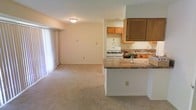 This single-floor residence offers a straightforward and functional layout centered around an open-concept living and kitchen area. The kitchen is equipped with warm-toned wooden cabinetry, a white gas range, a sink with a tiled backsplash, and a peninsula counter featuring a laminate surface that doubles as a casual dining or prep space. The peninsula creates a natural boundary between the kitchen and the adjoining living area, maintaining an open and connected feel throughout. The living area is finished with wall-to-wall carpeting in a neutral beige tone, contributing to a cozy and comfortable atmosphere. A large sliding glass door along one wall floods the space with natural light, while vertical blinds offer privacy and light control. The walls are painted in a soft, neutral tone that keeps the space feeling bright and airy. Recessed and flush-mount ceiling lighting ensures adequate illumination throughout. The overall design is practical and unpretentious, making efficient use of the available space while maintaining a welcoming residential ambiance suited to everyday living.