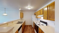 This single-floor residence offers a functional and comfortable layout with a cohesive design centered around warm oak wood tones and neutral color palettes throughout. The galley-style kitchen is a standout feature, lined on both sides with abundant honey-oak cabinetry providing generous storage. Light countertops run the length of the kitchen, complementing the wood-look flooring beneath. The kitchen is fully equipped with a coil-burner electric range, a refrigerator, a dishwasher, and a double-basin sink. A peninsula counter opens the kitchen toward the living area, enhancing the sense of connected space. The bedroom features soft carpeting underfoot, neutral walls, and a walk-in closet offering practical storage. A window brings in natural light and provides an outdoor view, while a baseboard heating unit ensures year-round comfort. The full bathroom is finished with hardwood-style flooring, an oak vanity with a single sink, a large mirror with wall-mounted lighting, and a shower-tub combination. The overall floor plan, visible in the dollhouse view, reflects a straightforward and efficient layout that balances open living areas with private spaces, all unified by consistent warm-toned finishes and practical design choices.