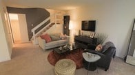 This two-story home offers a thoughtfully designed layout that balances comfort and style across both levels. The main living area serves as the heart of the space, featuring plush wall-to-wall carpeting and a warm, layered color palette. A bold dark-painted staircase accent wall creates a striking architectural focal point, complemented by crisp white stair railings that lead to the upper floor. The living room is furnished with a light gray sofa accented by terracotta and patterned throw pillows, arranged around a dark rectangular coffee table atop a rich rust-toned area rug. A woven pouf ottoman adds a natural texture element to the seating arrangement. Entertainment is centered around a wall-mounted flat-screen television positioned above a sleek dark media console, which provides both display and storage space. A tall arc floor lamp casts a warm ambient glow throughout the room, enhancing the cozy atmosphere. A dark shelving unit tucked beside the staircase offers additional decorative storage. The overall design aesthetic is modern and eclectic, blending dark furniture tones with warm neutrals and earthy accent colors to create a welcoming and well-curated living environment across both floors.