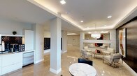 This single-floor space presents a polished and upscale lobby or common area distinguished by high-end finishes and thoughtful design throughout. The flooring consists of large-format beige marble tiles with a high-gloss finish, reflecting the ambient lighting and contributing to the space's bright, open feel. Structural columns with clean white bases punctuate the open floor plan, adding architectural definition without interrupting the sense of spaciousness.

The ceiling features a coffered tray design with recessed lighting, supplemented by a striking crystal chandelier that anchors the lounge area and serves as a central focal point. The lounge itself is furnished with contemporary white seating arrangements centered around a sleek glass coffee table adorned with floral accents, creating an inviting gathering area. A marble-clad accent wall with a decorative element adds a refined backdrop to this seating zone, and a large mirror enhances depth and light.

Toward one side of the space, a built-in mailbox unit provides practical functionality. A marble-topped round table with black chairs offers an additional seating option. The overall ambiance is sophisticated and well-lit, combining luxurious materials with modern design elements for an upscale communal environment.