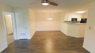 This single-floor space offers an open-concept layout that seamlessly connects the living area, dining space, and kitchen. The living area is equipped with a ceiling fan featuring an integrated light fixture, providing both comfort and functionality. Dark hardwood flooring runs throughout, adding warmth and a polished aesthetic to the interior.

The dining area is defined by an elegant chandelier with multiple arms, providing soft, ambient lighting overhead. The neutral, cream-colored walls complement the flooring and contribute to a bright, clean atmosphere throughout the space.

Adjacent to the dining area, the kitchen is partially visible and features white upper cabinetry, a decorative mosaic tile backsplash, and a built-in microwave. A raised counter separates the kitchen from the main living and dining areas, creating a natural division while maintaining the open feel.

A louvered closet door is situated near the living area, offering convenient storage access. The overall layout is straightforward and functional, with clean lines, consistent finishes, and a cohesive color palette that gives the space a move-in-ready appearance.