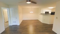 This single-floor space offers an open-concept layout that seamlessly connects the living area, dining space, and kitchen. The living area is equipped with a ceiling fan featuring an integrated light fixture, providing both comfort and functionality. Dark hardwood flooring runs throughout, adding warmth and a polished aesthetic to the interior.

The dining area is defined by an elegant chandelier with multiple arms, providing soft, ambient lighting overhead. The neutral, cream-colored walls complement the flooring and contribute to a bright, clean atmosphere throughout the space.

Adjacent to the dining area, the kitchen is partially visible and features white upper cabinetry, a decorative mosaic tile backsplash, and a built-in microwave. A raised counter separates the kitchen from the main living and dining areas, creating a natural division while maintaining the open feel.

A louvered closet door is situated near the living area, offering convenient storage access. The overall layout is straightforward and functional, with clean lines, consistent finishes, and a cohesive color palette that gives the space a move-in-ready appearance.