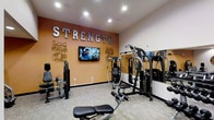 This fitness facility is designed to support a comprehensive strength training regimen in a focused and motivating environment. The strength training area serves as the centerpiece of the space, featuring a curated selection of modern resistance and cable machines suited for a full-body workout. A bold, warm orange accent wall anchors the room, emblazoned with the word "STRENGTH" in large metallic lettering, reinforcing the purpose of the space with an energetic visual statement. Decorative geometric wall art flanks a wall-mounted flat-screen television, offering entertainment during workouts. The flooring consists of neutral, low-pile carpeting that provides a practical surface throughout the area. Recessed ceiling lighting ensures the space is well-lit and evenly illuminated. A full-length mirror runs along one wall, expanding the sense of space and allowing users to monitor their form during exercises. The overall ambiance is clean, purposeful, and energizing, with a color palette and layout designed to keep users motivated. The facility presents a well-organized and accessible environment for strength-focused training.