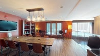 The WiFi Lounge is a thoughtfully designed communal space that blends functionality with a warm, inviting aesthetic. Bold terracotta-hued walls set an energetic tone throughout, complemented by wood-look flooring that adds a sense of warmth and cohesion. The space is organized into distinct seating zones to accommodate a variety of uses. A large, rustic wooden communal table anchors the central area, suspended beneath a striking linear pendant light fixture with multiple glass globe pendants, making it ideal for collaborative work or group gatherings. Surrounding the table are tan leather chairs that reinforce the lounge's earthy color palette.

Along the windowed wall, multiple high-top round tables paired with contemporary bar stools create a casual seating area bathed in natural light from shuttered French-style windows. Floor lamps provide additional ambient lighting throughout. A wall-mounted flat-screen TV is positioned prominently on one wall, accompanied by a pair of built-in wooden shelving units displaying decorative objects. Recessed lighting in the ceiling provides even overhead illumination. A glass partition wall on one side separates the lounge from an adjacent area, adding transparency while maintaining a defined, comfortable space for connectivity and productivity.