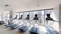 Located at 505 N State St in Chicago's River North neighborhood, this fitness center offers a well-equipped, modern workout facility with an inspiring urban backdrop. The space is defined by expansive floor-to-ceiling windows that frame sweeping panoramic views of the Chicago skyline, flooding the interior with natural light and creating an energizing atmosphere for exercise.

The cardio zone features a row of treadmills and elliptical machines positioned directly facing the city views, allowing users to exercise while taking in the surrounding high-rise landscape. A full wall of mirrors runs along one side of the main floor, visually expanding the space and enhancing the open, airy feel of the room.

The free weights area is equipped with a tiered dumbbell rack, a workout bench, and stability equipment including a BOSU ball, accompanied by framed artwork that adds a touch of sophistication to the environment. Additional equipment includes rowing machines, a cable machine, and stationary cycling bikes, providing a comprehensive range of training options.

The facility also includes a reception and check-in counter with a stainless steel surface, a water dispenser, and wood-paneled entry doors. The neutral-toned flooring, white walls, and clean architectural lines throughout contribute to a functional yet polished aesthetic. This fitness center is designed to serve the needs of dedicated exercisers within a professional, well-maintained environment.