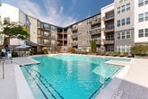 Located at 747 Ralph McGill Blvd NE in Atlanta, Georgia, this outdoor amenity space is centered around a generously sized resort-style swimming pool with crystal-clear turquoise water. The pool is framed by a spacious concrete sundeck lined with wicker chaise lounges and reclining chairs, providing ample room for relaxation. Manicured landscaping, including tall evergreen trees, flowering shrubs, and raised garden planters, adds greenery and visual interest throughout the courtyard. A dedicated outdoor dining area features a table with seating and a striped umbrella, ideal for al fresco gatherings. The courtyard is enclosed by a sleek, black iron security fence with gated access, ensuring privacy and safety. An outdoor shower station is conveniently positioned near the pool area. The space is surrounded on multiple sides by a contemporary multi-story building with a modern exterior of gray and dark blue paneling, large windows, and private balconies on each level that overlook the courtyard below. Warm wood-clad ceiling accents are visible on select balcony overhangs, adding an architectural detail to the facade. The open-sky setting allows abundant natural light to fill the courtyard throughout the day, creating a bright and welcoming outdoor environment well-suited for leisure and community life.