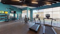 Located at 4625 Millbrook Green Dr in Raleigh, NC, this well-equipped fitness center offers a dynamic and energizing environment designed to support a wide range of workout routines. The space features a distinctive split-level layout, with the upper level housing a row of cardio machines — including treadmills and ellipticals — and the lower level providing an open area ideal for functional training, complete with a punching bag, battle ropes, and a flat bench. Bold teal accent walls contrast with crisp white surfaces, lending the space a modern and motivating aesthetic. Exposed dark ceiling beams, ceiling fans, and linear lighting fixtures contribute to the stylish yet functional interior design. Floor-to-ceiling glass windows and wall-length glass partitions flood the space with abundant natural light and offer views of the surrounding community. Rubberized flooring throughout provides safety and comfort underfoot. A dedicated strength training area features a cable machine and a full dumbbell rack with a wall-mounted mirror. An adjacent stretch and mat area is equipped with a mounted Smart Mat core exercise guide, offering additional space for warm-ups and cooldowns. A wall-mounted television adds entertainment convenience during workouts.