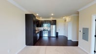 This single-floor residence offers a well-designed open-concept layout that seamlessly connects the kitchen and living area. The kitchen is a standout feature, showcasing rich, dark espresso-toned cabinetry that extends from floor to ceiling, providing generous storage capacity. Stainless steel appliances, including a refrigerator, electric range, and microwave, lend a modern, polished look to the space. A built-in sink with additional cabinetry completes the functional kitchen setup. The flooring transitions from deep, dark hardwood in the kitchen and main living area to plush carpeting, adding warmth and comfort underfoot. Crown molding lines the ceiling throughout, adding an elegant architectural detail that elevates the overall aesthetic. Recessed lighting is thoughtfully placed to illuminate the space evenly, while a ceiling-mounted light fixture provides additional brightness. A set of white panel doors — including a spacious closet with transom detail above — anchors the entryway area, offering practical storage solutions. The light-toned walls contrast beautifully with the dark cabinetry and flooring, creating a balanced and inviting ambiance throughout this single-level home.