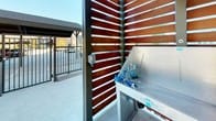 This outdoor dog park and wash area is a well-appointed pet amenity space designed to serve both residents and their four-legged companions. The facility is anchored by a professional-grade stainless steel bathing tub, branded by Groomer's Best, equipped with a built-in hose and faucet system for convenient pet washing. A stainless steel grooming table with adjustable arm support is also available within the enclosure, providing a dedicated space for post-bath grooming and drying. The wash station features a step entry for easy pet access.

The enclosure is constructed with striking horizontal wood slat privacy walls set within a steel frame, creating a modern aesthetic while allowing natural light and airflow to filter through. The slatted design casts dynamic light patterns throughout the day, adding visual interest to the functional space. A gated entry with a secure latch provides controlled access to the wash station.

Surrounding the enclosure is a generously sized open-air dog park featuring well-maintained green lawn, decorative rock borders, and young shade trees. Metal perimeter fencing with a gated entry defines the park area, ensuring a safe environment for pets to roam. The overall design balances practicality with a clean, contemporary aesthetic that complements the modern residential building backdrop.