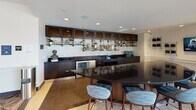 Located at 901 H St NE in Washington, DC's vibrant NoMa/H Street corridor, this thoughtfully designed amenity floor offers a diverse range of social and recreational spaces unified by a bold, eclectic aesthetic. The entry lobby commands attention with its intricate geometric parquet flooring, a sweeping gold-accented ceiling detail, and clusters of emerald green velvet seating beneath dramatic pendant lights—setting a sophisticated tone throughout.

The lounge area features a plush sectional sofa, a large built-in media wall flanked by dark wood cabinetry, and expansive windows that flood the space with natural light and neighborhood views. A dedicated dining area is anchored by an oval table with artistic pendant fixtures above, and floor-to-ceiling sliding glass doors that open directly onto an outdoor terrace complete with lounge furniture.

A standout fireplace nook showcases a stone surround with curated book storage, framed by large windows on either side offering terrace views—creating an inviting retreat. The full-service bar area presents a sleek black granite countertop, dark cabinetry, industrial-style open shelving, and bar seating, ideal for social gatherings. A game room with arcade machines and additional seating rounds out the offerings, ensuring a well-appointed space for both relaxation and entertainment.