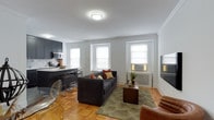 This single-floor residence combines classic architectural details with modern updates to create a comfortable and stylish living environment. The open-concept living and dining area is anchored by warm parquet wood flooring, crown molding, and multiple windows that fill the space with natural light. A large sectional sofa, coffee table, and wall-mounted television make this a functional and inviting gathering space.

The kitchen has been thoughtfully renovated with dark shaker-style cabinetry, gold hardware, stainless steel appliances including a refrigerator, range, microwave, and dishwasher, complemented by a herringbone tile backsplash and crisp white countertops. A center island provides additional prep and seating space.

The residence features multiple well-proportioned bedrooms, each finished with the same classic parquet flooring and crown molding. The rooms are bright, with windows fitted with blinds, and offer ample space for full-sized furniture arrangements.

The bathroom stands out with floor-to-ceiling marble-style tile, a soaking tub, and a sleek stainless steel multi-function shower panel, delivering a spa-like experience. Clean white cabinetry completes the refined look. Throughout the residence, the consistent use of neutral tones and quality finishes creates a cohesive and polished ambiance.