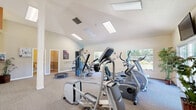 Located at 996 Meridian Ave in San Jose, CA, this fitness center offers a well-equipped, single-floor layout designed to support a variety of exercise and recreational activities. The centerpiece of the facility is a generously sized exercise room featuring vaulted ceilings that soar overhead, bathed in natural light from multiple skylights and large windows that frame views of surrounding greenery. The open, carpeted floor plan accommodates a wide selection of cardio and strength equipment, including treadmills, elliptical trainers, a stationary bike, and a stair climber, as well as free weights and a trampoline. A ping-pong table occupies a dedicated area of the room, adding a recreational dimension to the space. Decorative artwork with fitness-themed motifs adorns the walls, contributing to a motivating atmosphere. A wall-mounted television and a wall clock are also present for added convenience. Two restrooms serve the facility, each finished with two-tone painted walls, wainscoting, and durable tile flooring in a diamond pattern. One restroom includes a shower stall, offering added functionality for users. The overall ambiance is bright, airy, and well-maintained, making this fitness center a practical and inviting space for daily exercise.