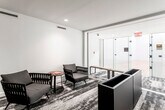 This single-floor recreational facility is thoughtfully designed around a regulation racquetball court, combining athletic functionality with a refined, modern aesthetic. The court itself is the centerpiece of the space, featuring a polished light maple hardwood floor with crisp red boundary lines clearly marked throughout. Soaring white walls extend to impressive heights on all sides, creating an expansive, open playing environment. Overhead, linear fluorescent lighting provides even, bright illumination across the entire court surface. Entry to the court is managed through full-height glass panel doors fitted with sturdy hardware, allowing clear visibility between the court and the adjacent lounge area.

The lounge area adjoining the court offers a welcoming place for players to prepare or unwind. It features a patterned carpet with abstract tones of grey and taupe, providing a stylish contrast to the crisp white walls. Contemporary woven armchairs in warm grey tones are arranged in casual groupings alongside a round wood accent table. Recessed ceiling lighting keeps the area well-lit and polished. A keyed-access door within the lounge leads to additional facilities. Together, the court and lounge create a cohesive, well-appointed space suited for both serious play and comfortable relaxation.
