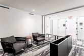 This single-floor recreational facility is thoughtfully designed around a regulation racquetball court, combining athletic functionality with a refined, modern aesthetic. The court itself is the centerpiece of the space, featuring a polished light maple hardwood floor with crisp red boundary lines clearly marked throughout. Soaring white walls extend to impressive heights on all sides, creating an expansive, open playing environment. Overhead, linear fluorescent lighting provides even, bright illumination across the entire court surface. Entry to the court is managed through full-height glass panel doors fitted with sturdy hardware, allowing clear visibility between the court and the adjacent lounge area.

The lounge area adjoining the court offers a welcoming place for players to prepare or unwind. It features a patterned carpet with abstract tones of grey and taupe, providing a stylish contrast to the crisp white walls. Contemporary woven armchairs in warm grey tones are arranged in casual groupings alongside a round wood accent table. Recessed ceiling lighting keeps the area well-lit and polished. A keyed-access door within the lounge leads to additional facilities. Together, the court and lounge create a cohesive, well-appointed space suited for both serious play and comfortable relaxation.
