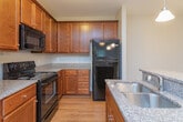 Located at 102 Village Dr in Mebane, North Carolina, this single-floor residence offers a thoughtfully designed layout with functional living spaces throughout. The kitchen is a standout feature, showcasing rich warm-toned wood cabinetry, granite-style countertops, and black appliances including a range, microwave, refrigerator, and dishwasher. A peninsula with an integrated sink and pendant lighting above provides both additional prep space and a casual dining option, while hardwood-style flooring adds warmth underfoot. The living areas are finished with neutral-toned carpet and ceiling fans, creating a comfortable and cohesive atmosphere. French doors in the main living space open to the outdoors, inviting natural light and providing exterior access. The bedroom is generously sized, featuring multiple windows, a ceiling fan, and soft carpeting, with access to a spacious walk-in closet outfitted with wire shelving and hanging rods along multiple walls. The bathroom is finished with tile flooring, a wood vanity with granite-style countertop, a large mirror, and a tub-shower combination. A dedicated laundry area with washer and dryer connections, overhead shelving, and tile flooring adds everyday convenience. Clean lines, neutral tones, and consistent finishes unify the space throughout.